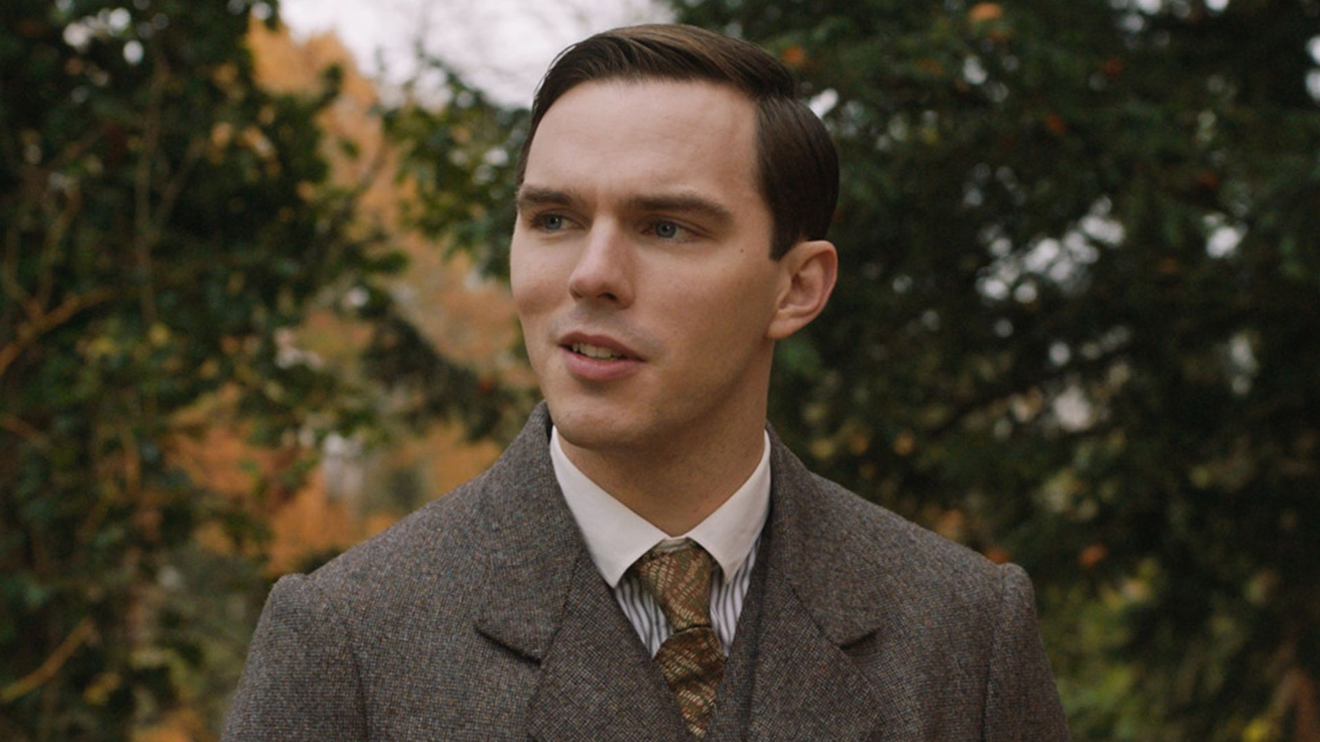 Nicholas Hoult de fato castanho e gravata, de pé na rua no papel do jovem J.R.R. Tolkien