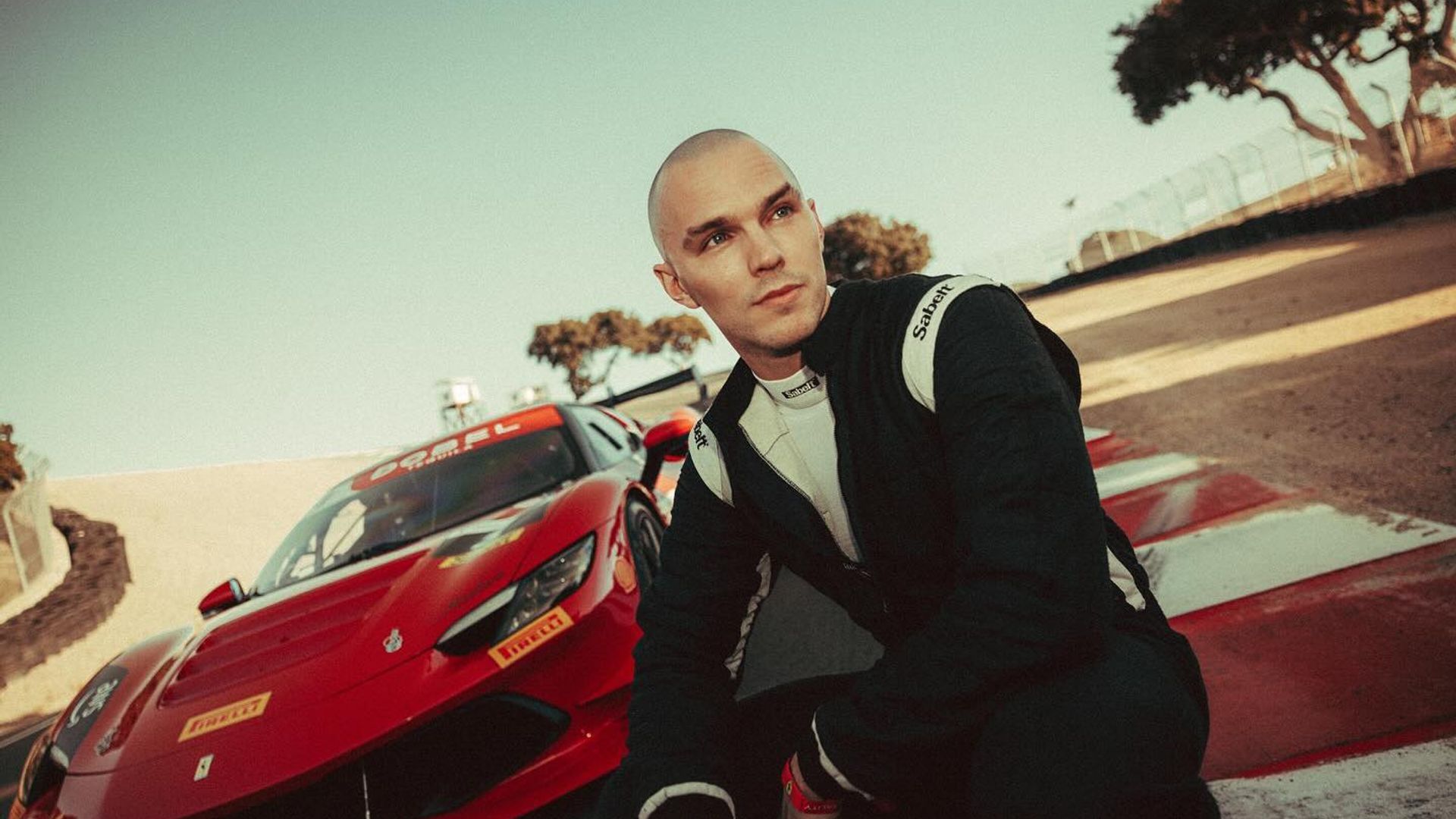 Nicholas Hoult num fato de corrida sentado na pista com um carro desportivo Ferrari vermelho atrás dele