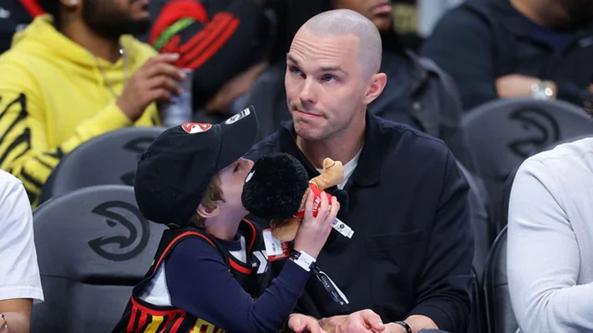 Nicholas Hoult sentado nas bancadas num jogo de basquetebol ao lado do seu filho que segura um brinquedo