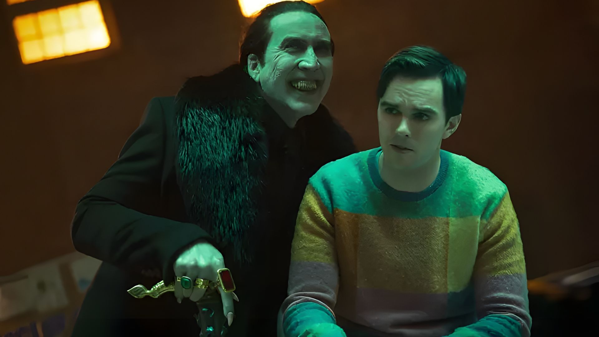 Nicolas Cage com roupas pretas e anéis paira ameaçadoramente sobre Nicholas Hoult numa camisola colorida