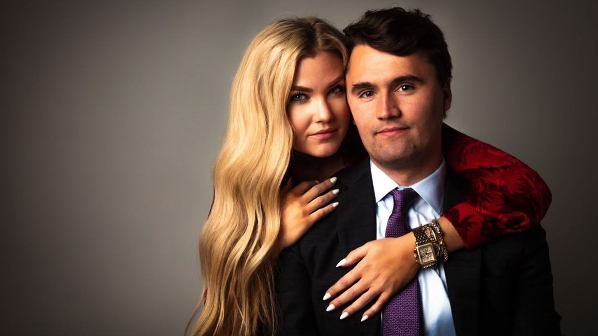 Charlie Kirk e Erica Frantzve abraçam-se em frente à câmara contra um fundo escuro