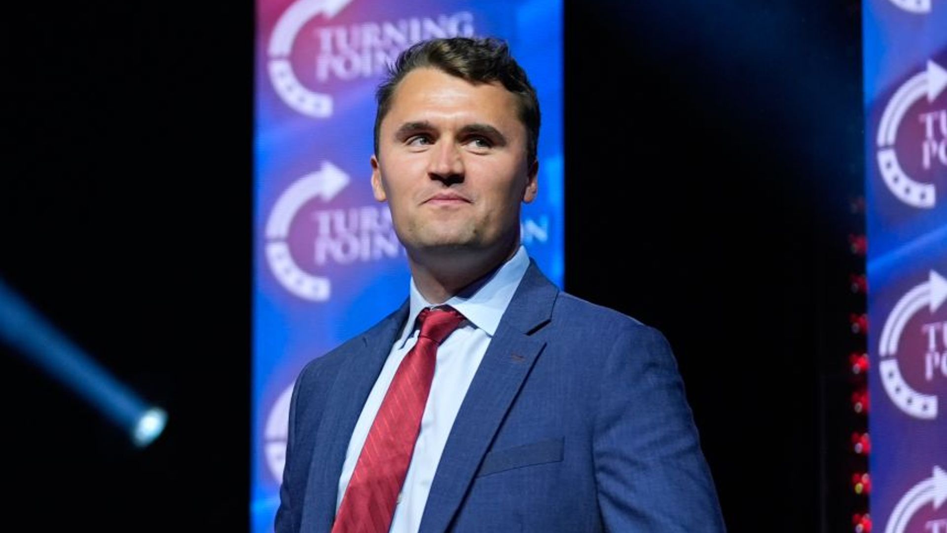 Charlie Kirk posa orgulhosamente contra um fundo escuro vestindo um fato azul