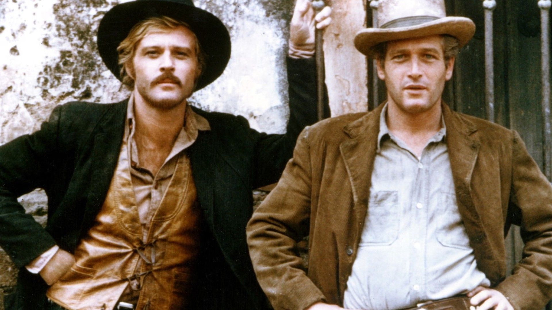 Robert Redford e Paul Newman com chapéus de cowboy e casacos, lado a lado junto a uma parede, posando para uma fotografia