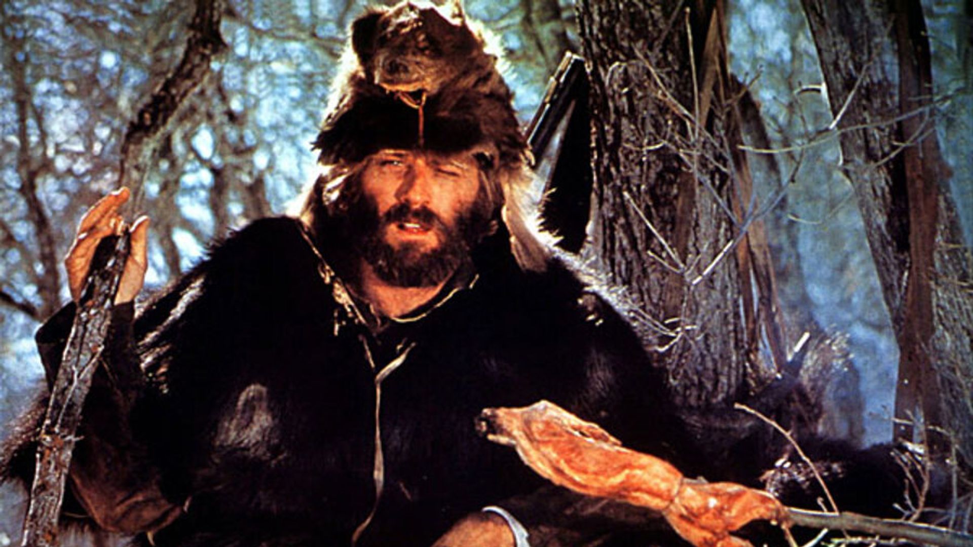 Robert Redford vestido em peles e com um gorro feito de cabeça de animal, grelhando carne numa fogueira na floresta, cena do filme "Jeremiah Johnson"