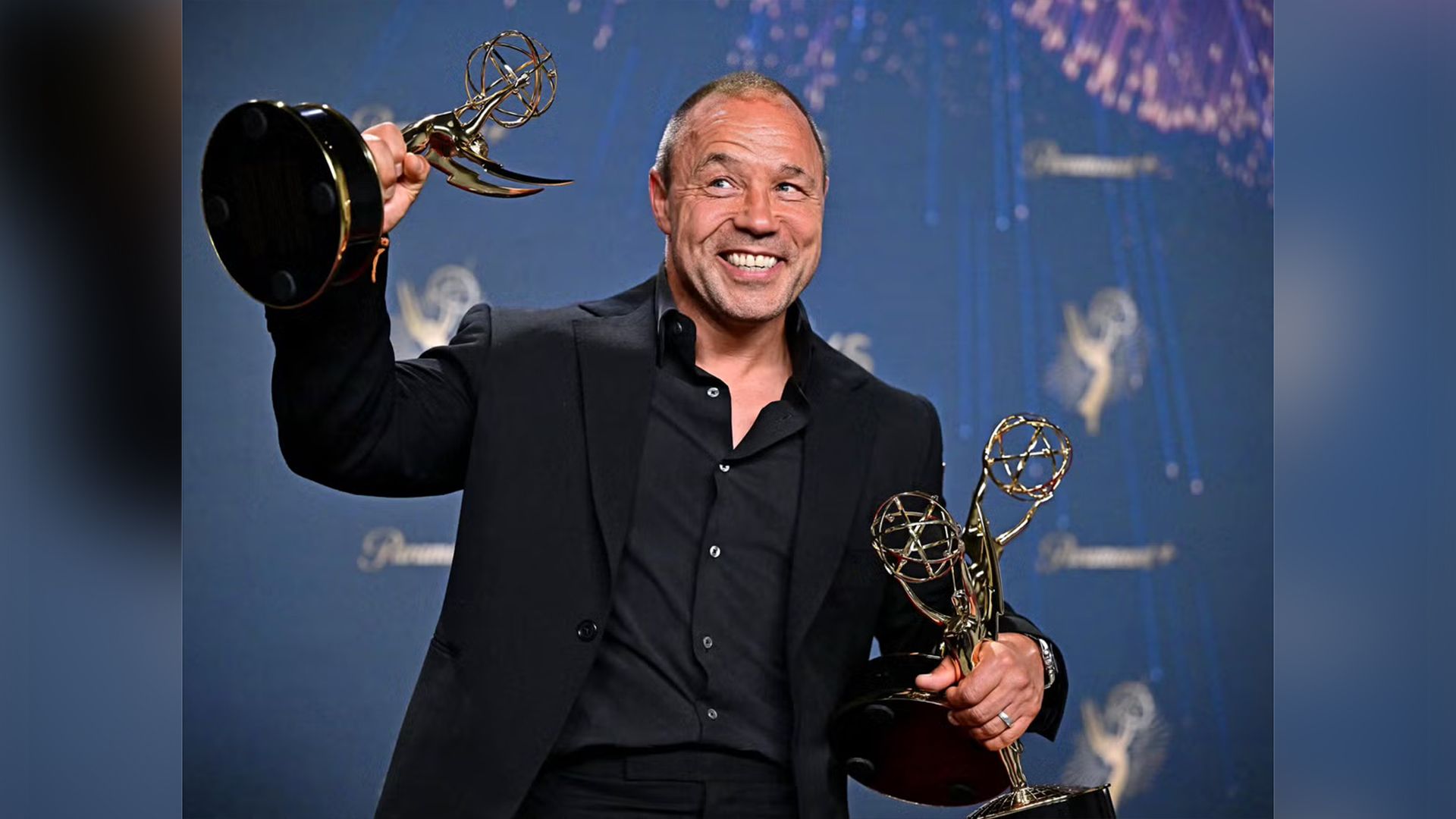 Stephen Graham de fato preto segura três estatuetas douradas dos Emmy nas mãos, sorrindo amplamente