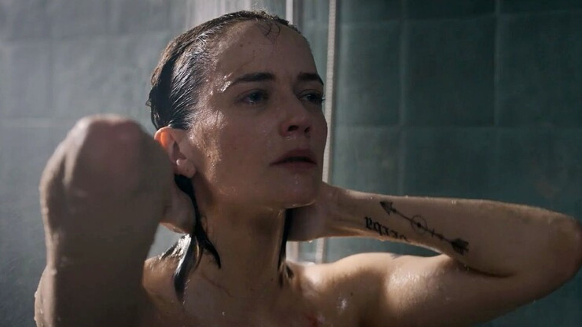 Eva Green com cabelo molhado a tomar duche, mãos atrás da cabeça, apenas o rosto e braço tatuado visíveis