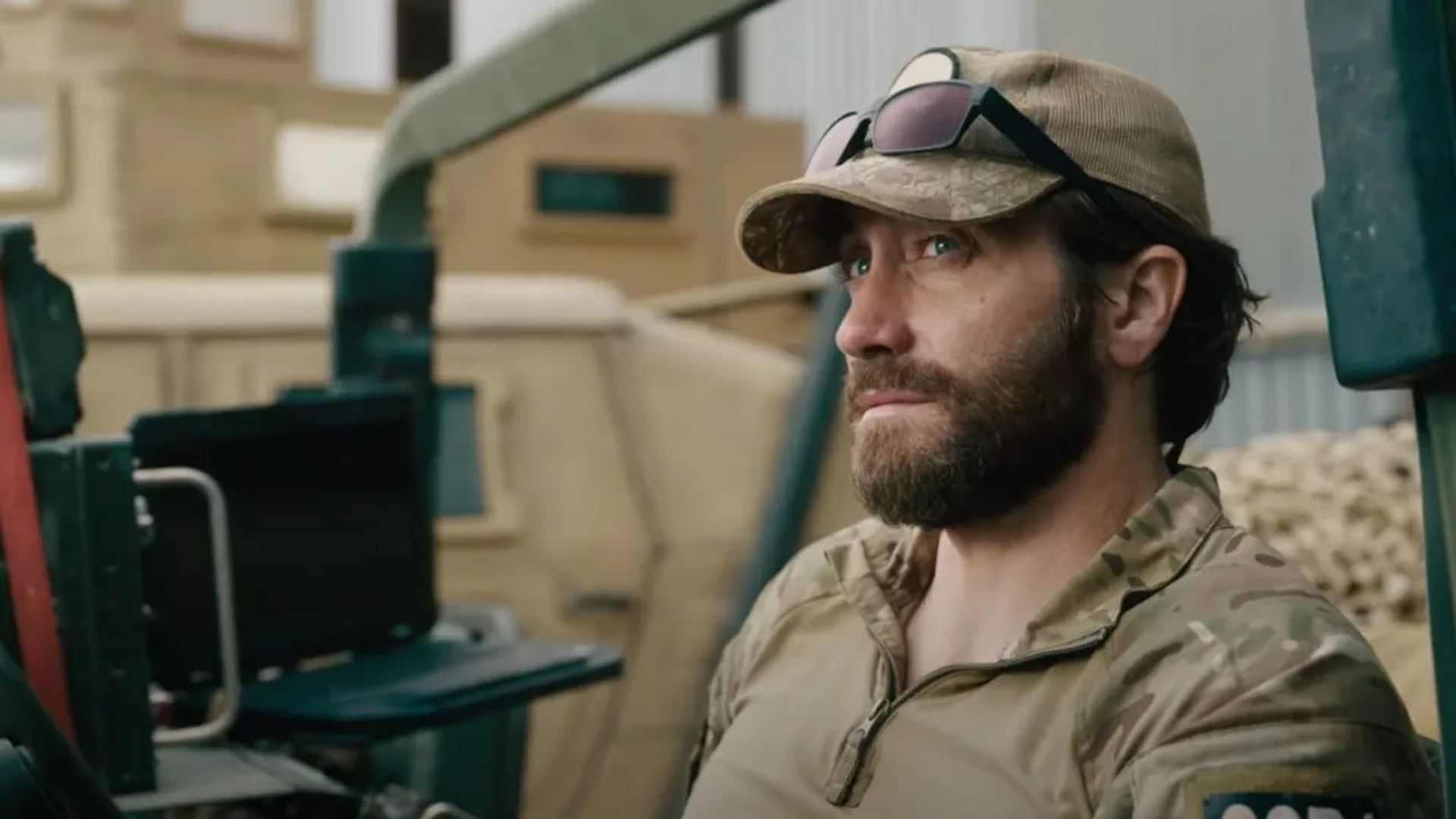 Jake Gyllenhaal em uniforme militar e boné com óculos na pala sentado olhando para o lado com expressão séria