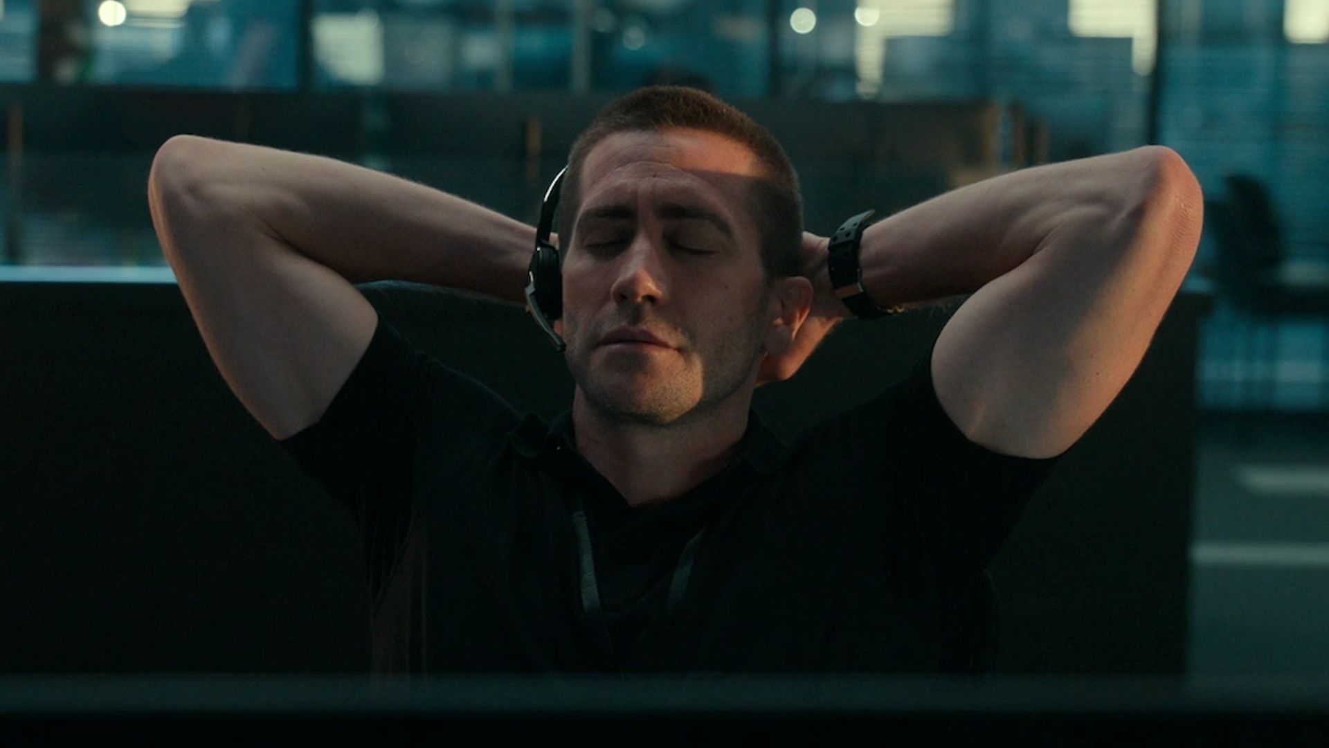Jake Gyllenhaal usando fones de ouvido com microfone sentado com os olhos fechados, mãos atrás da cabeça