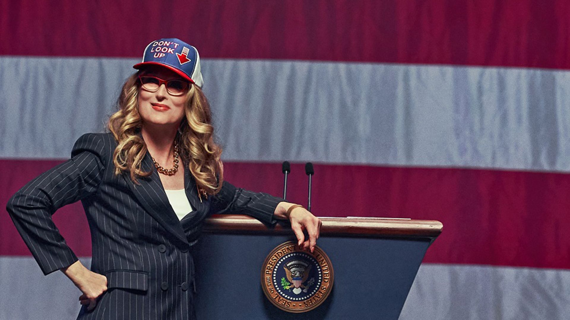 Meryl Streep de fato e boné com bandeira americana está contra um cenário da bandeira dos EUA, encostada a um pódio