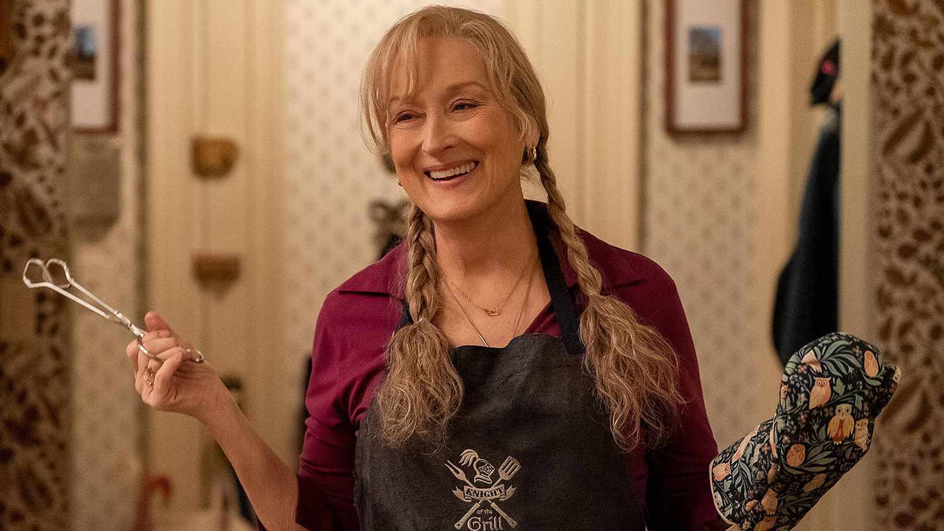 Meryl Streep com tranças segura uma pinça numa mão, usando uma luva de cozinha na outra