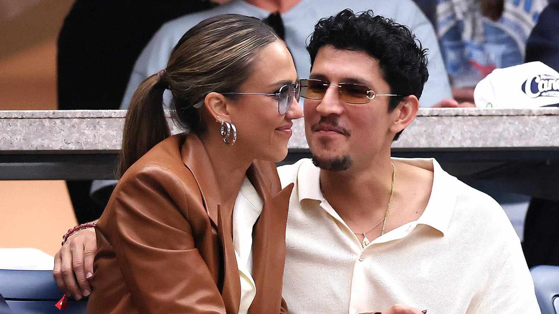Jessica Alba e Danny Ramirez sentados nas bancadas de um evento desportivo abraçados