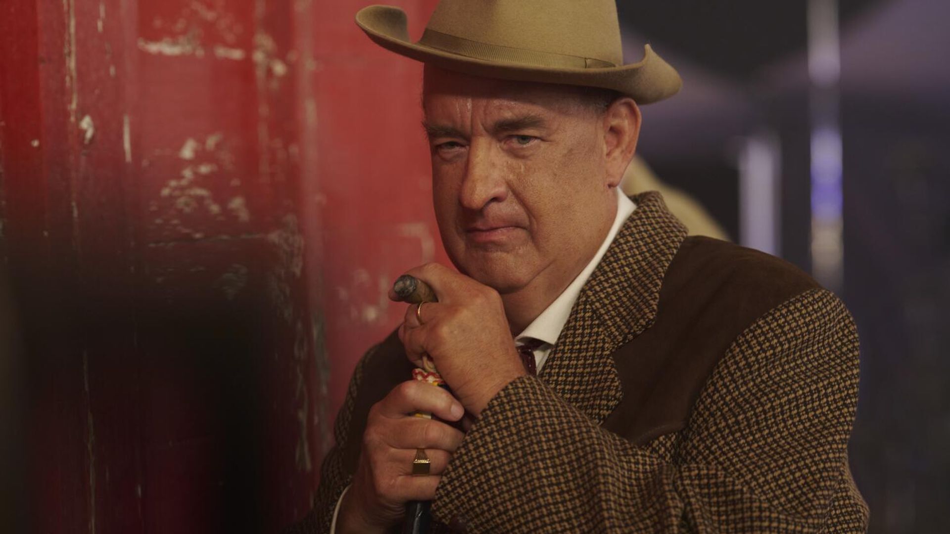 Tom Hanks em fato castanho e chapéu apoia-se numa bengala, olhando seriamente para a câmara