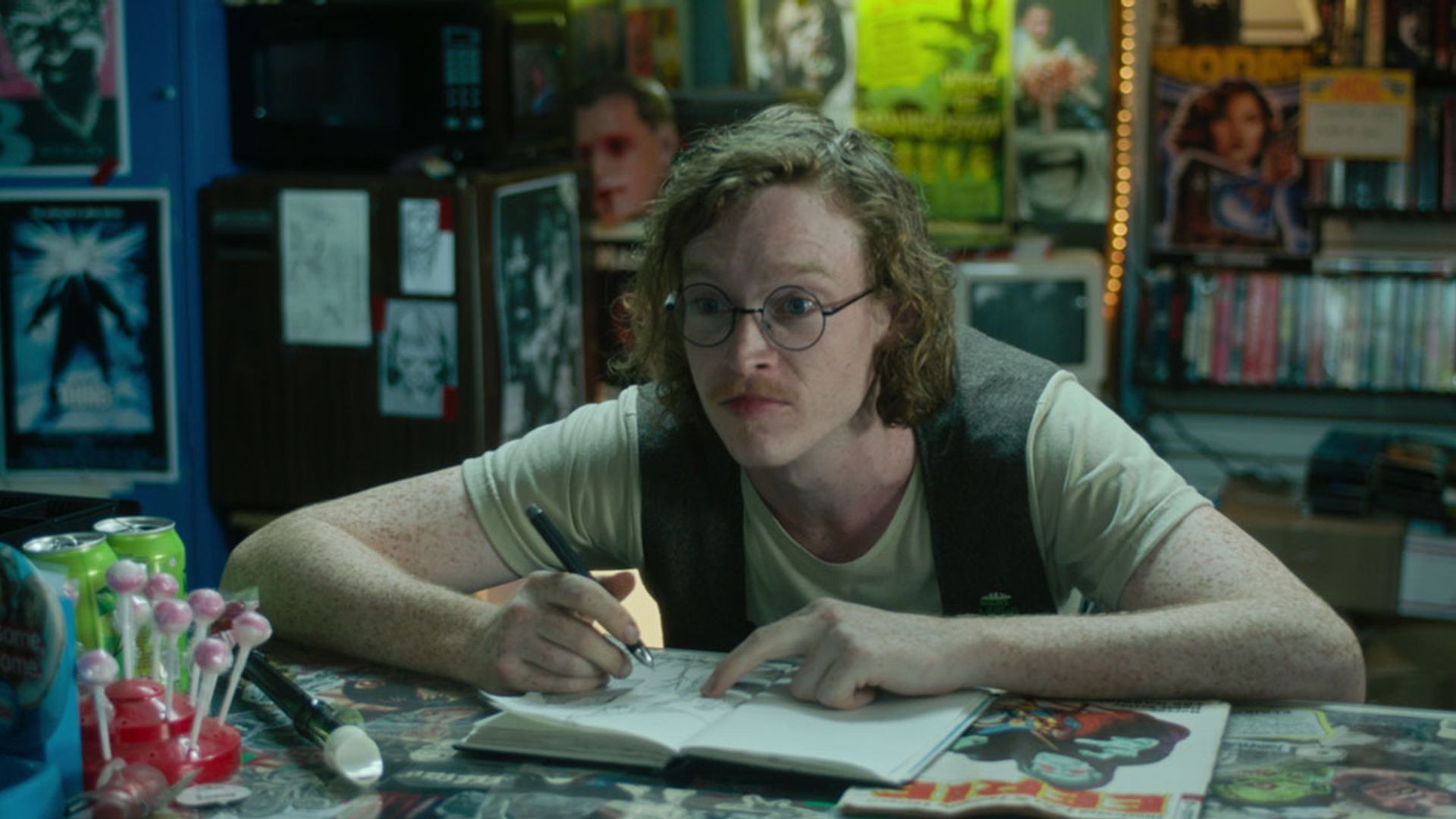 Caleb Landry Jones usando óculos e colete senta-se numa mesa com um bloco de notas, desenhando e olhando para o lado