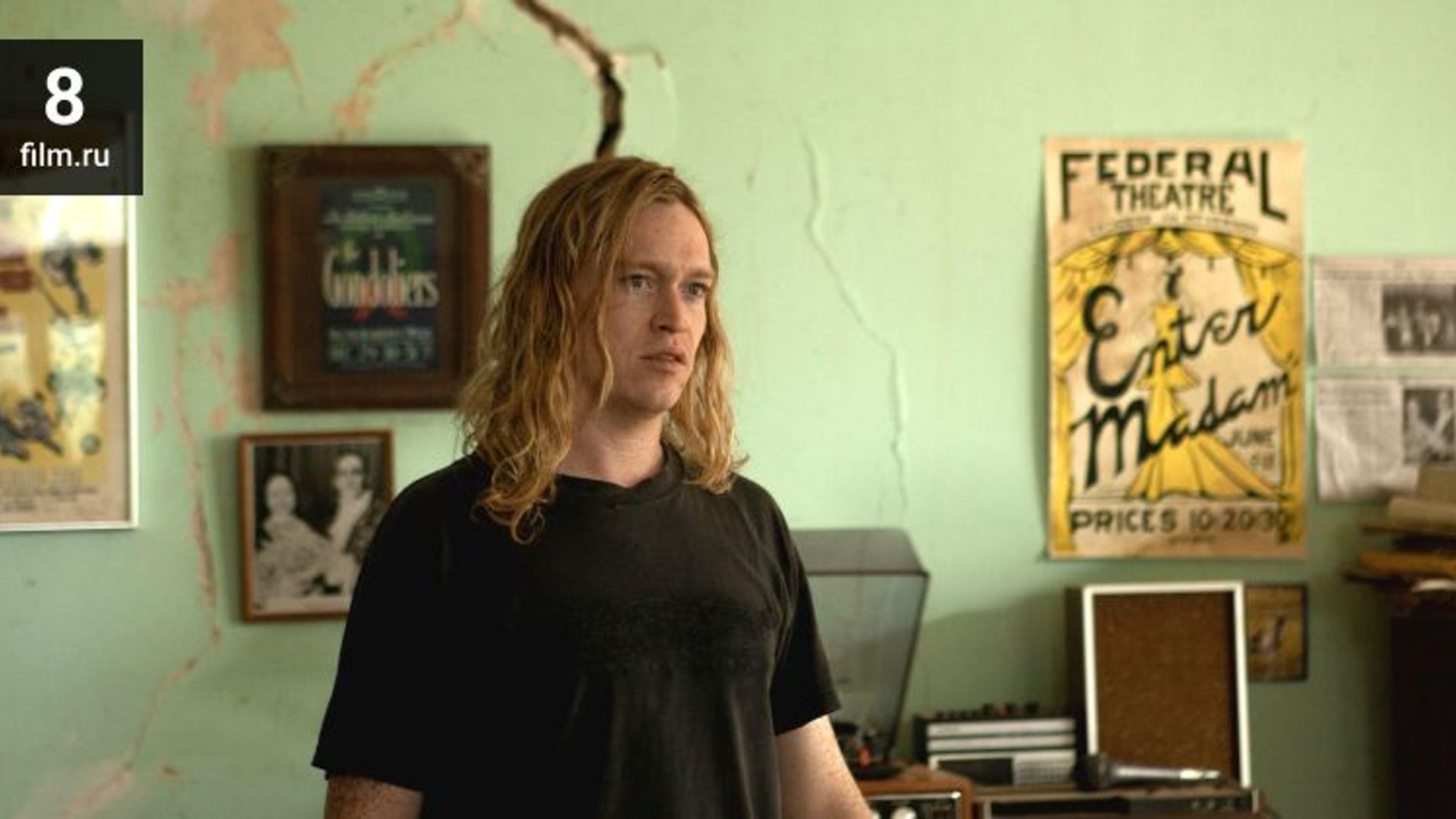 Caleb Landry Jones com cabelo loiro comprido numa t-shirt preta está numa sala com posters na parede