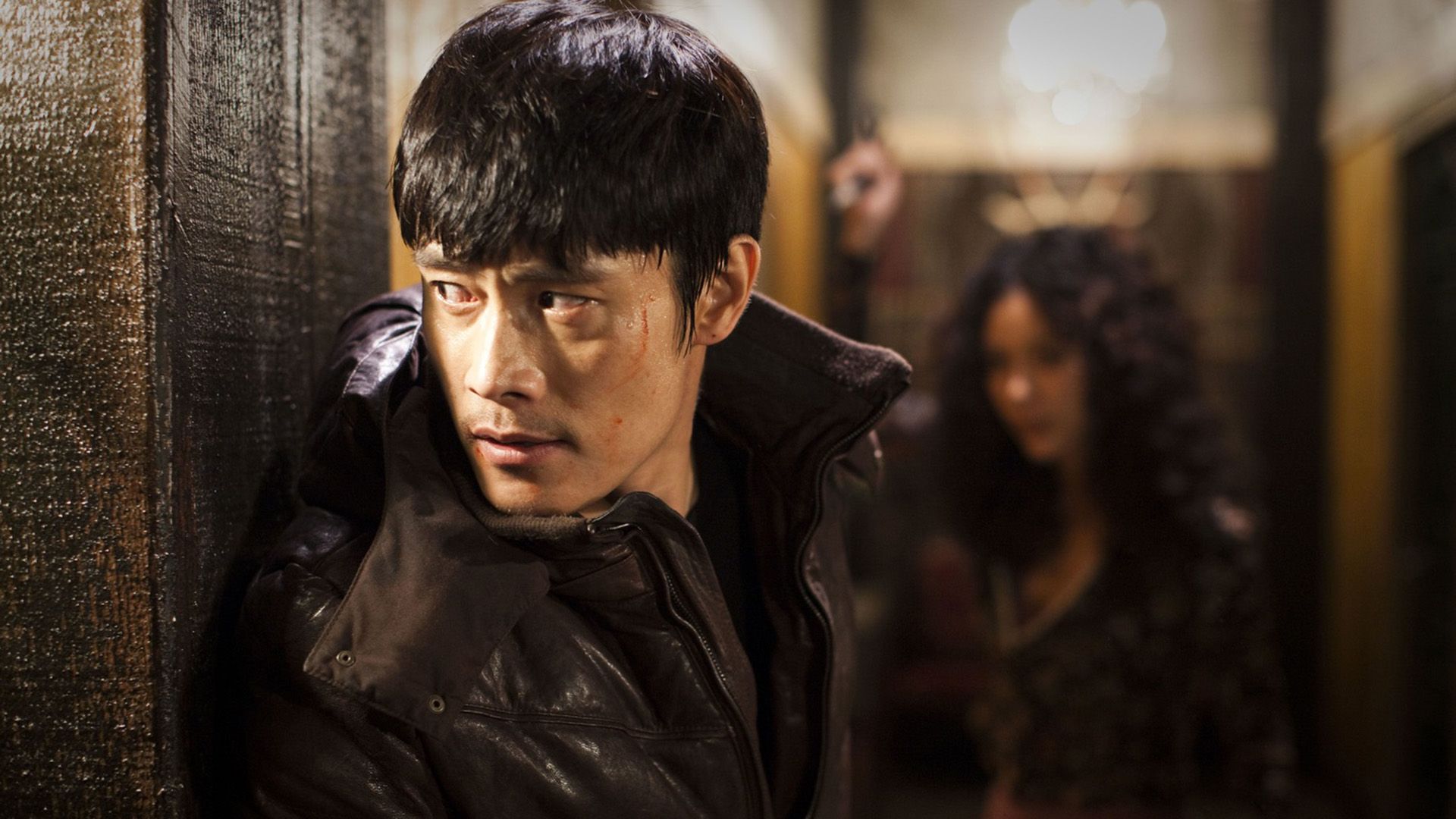Lee Byung-hun, de casaco preto, espreita ao virar da esquina de um edifício