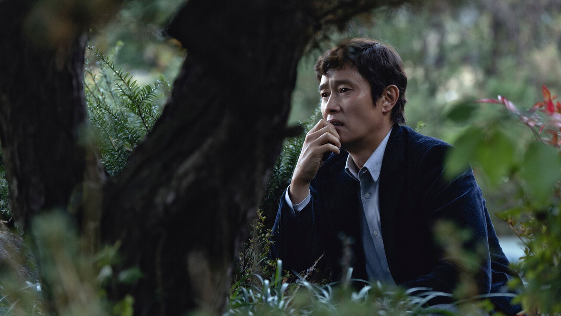 Lee Byung-hun com um blazer azul sentado na relva entre árvores, com desespero estampado no rosto