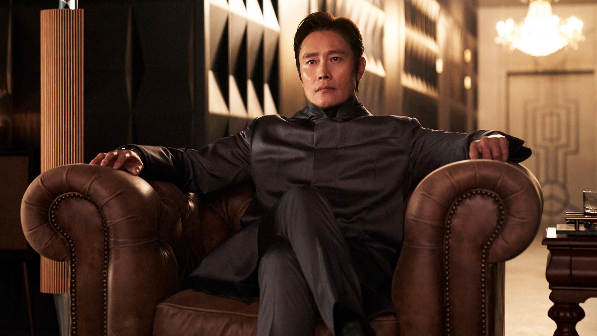 Lee Byung-hun num blazer cinzento sentado numa poltrona de couro com as pernas cruzadas