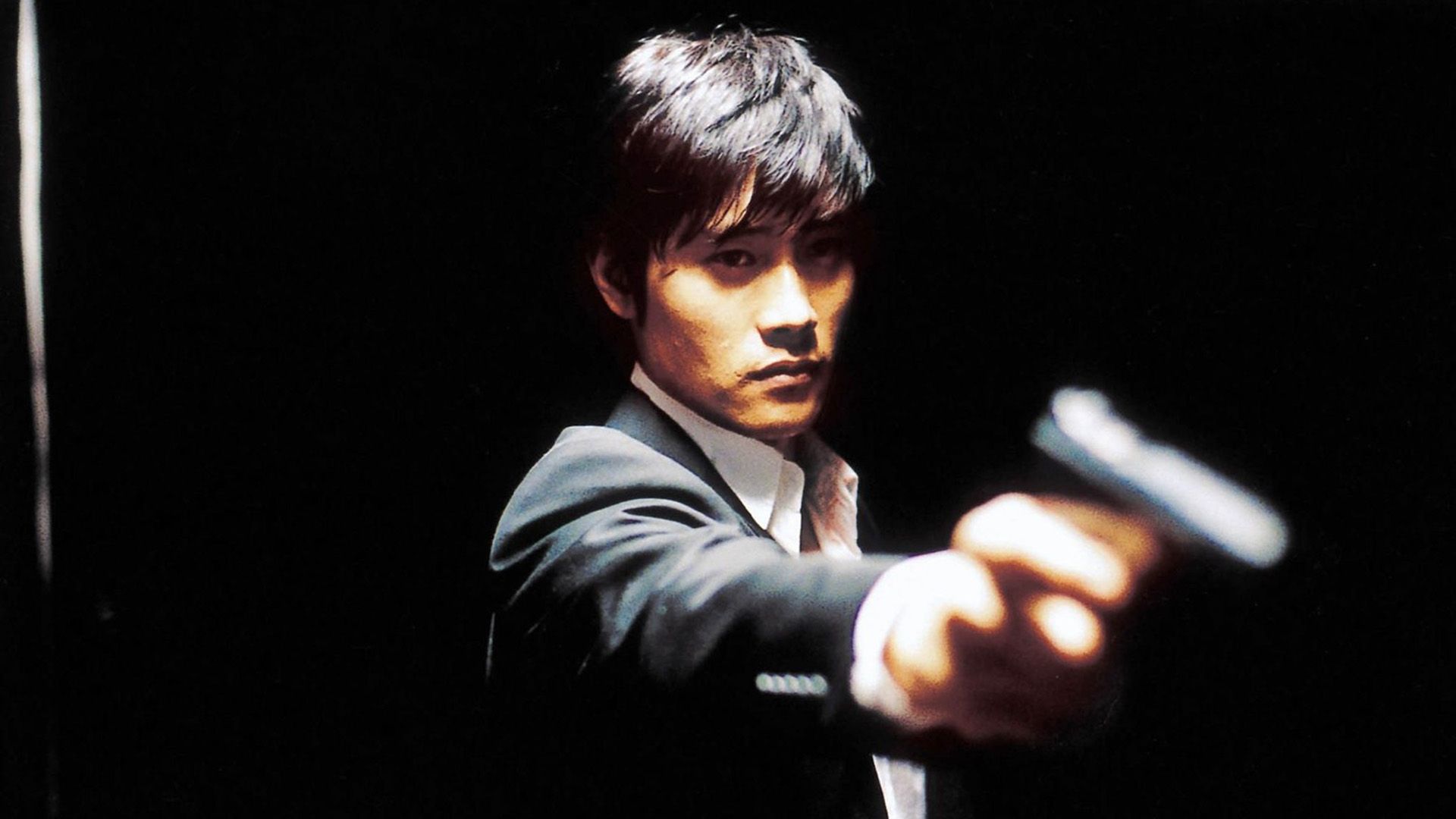Lee Byung-hun, de camisa branca e casaco, segura uma arma com o braço estendido