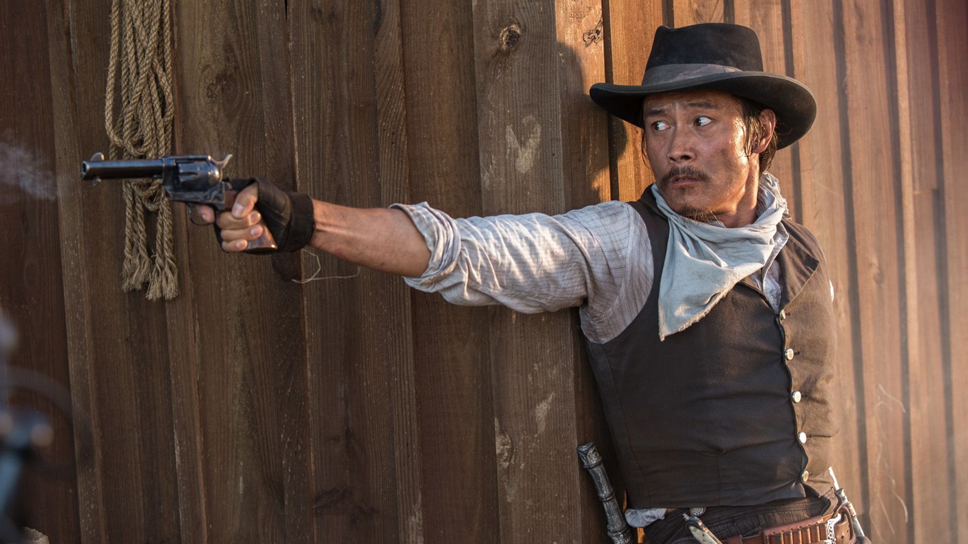 Lee Byung-hun, vestido de cowboy e com chapéu, dispara uma pistola
