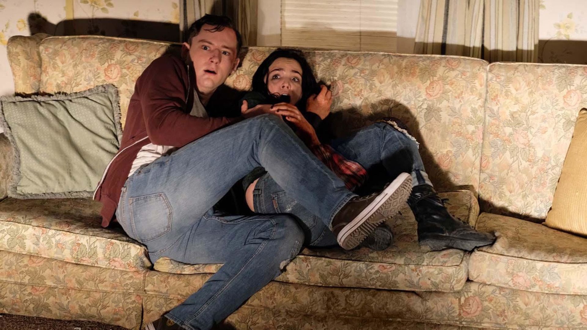 Lewis Pullman e uma rapariga sentados aterrorizados num sofá com as pernas encolhidas