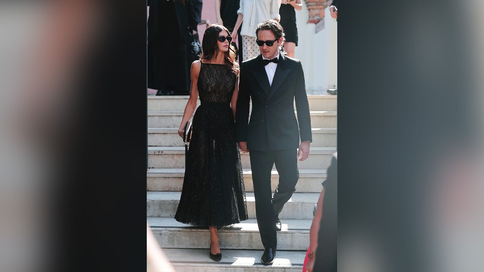 Lewis Pullman de smoking preto e Kaia Gerber com vestido de noite preto descem as escadas, ambos usando óculos escuros