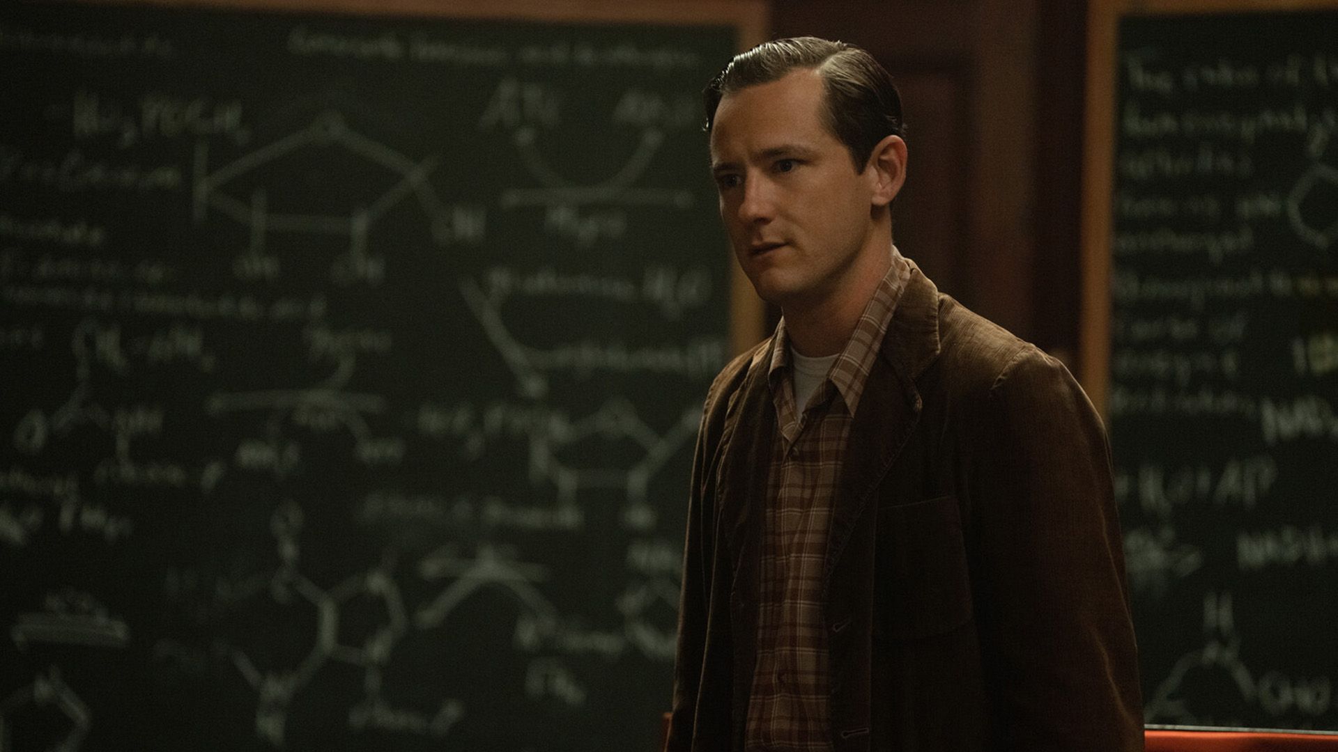 Lewis Pullman num blazer castanho de bombazina e camisa aos quadrados está encostado a um quadro com fórmulas químicas