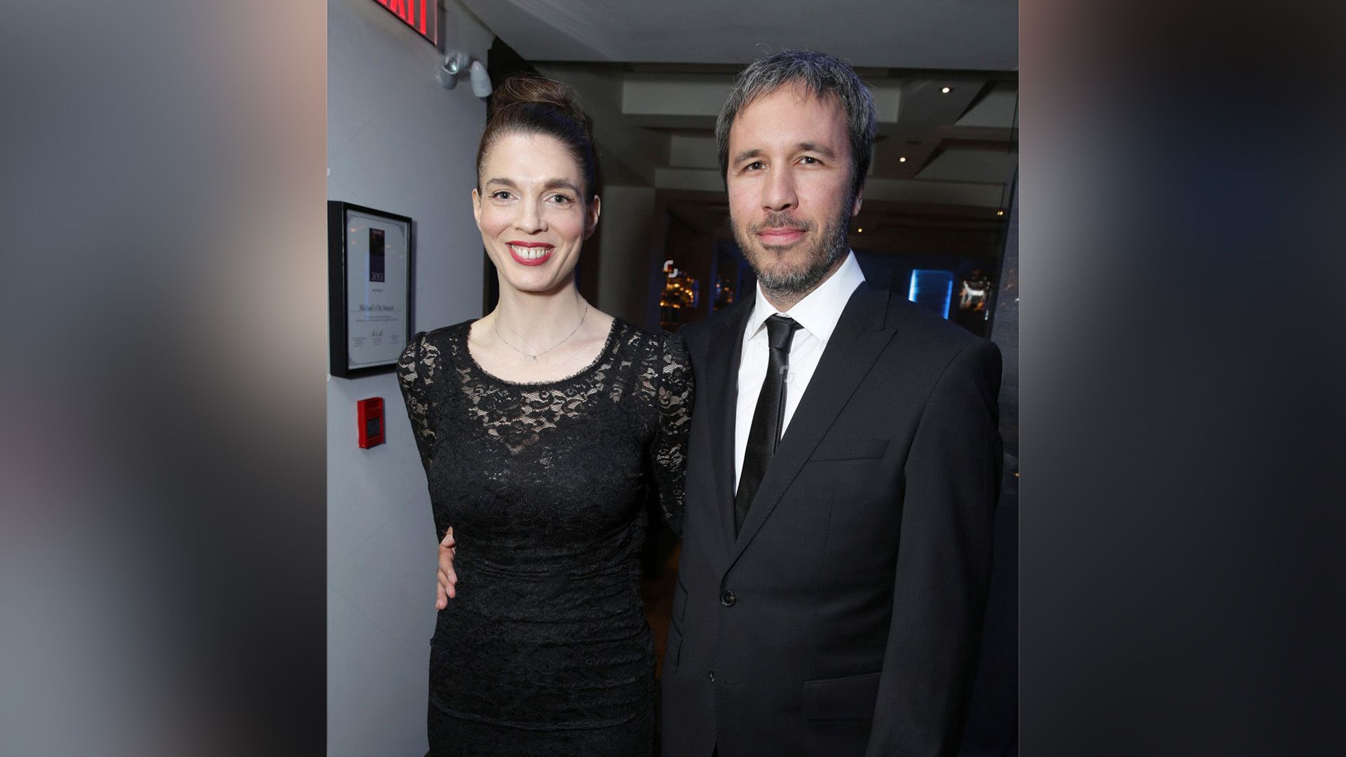 Denis Villeneuve de fato abraça Masha Grenon sorridente num vestido preto de renda pela cintura