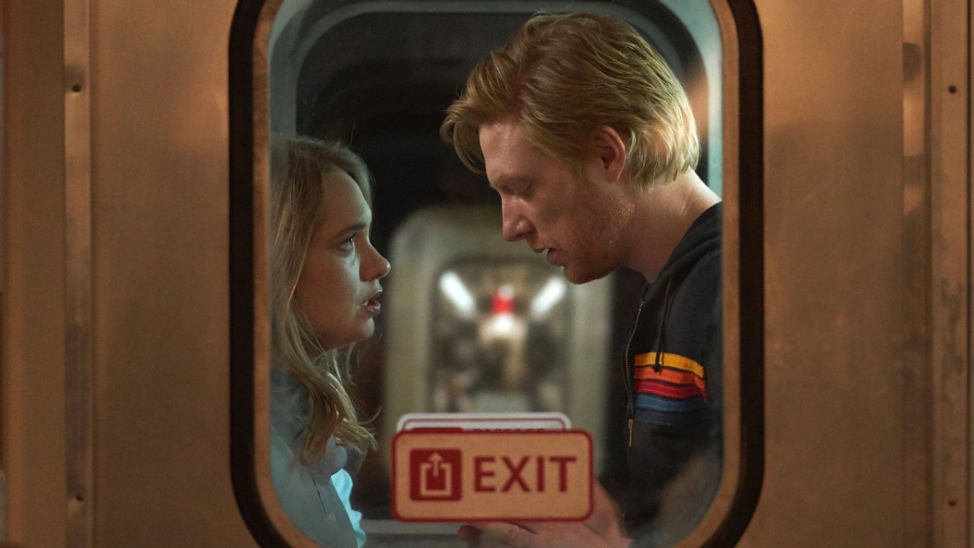Domhnall Gleeson e Merritt Wever frente a frente no vestíbulo de um comboio, filmados do exterior através da janela