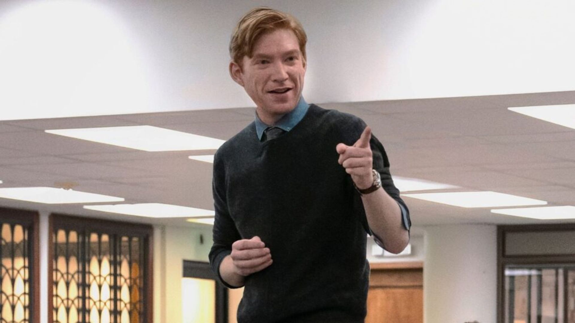 Domhnall Gleeson numa camisola azul-escura em pé sobre uma mesa e a apontar o dedo