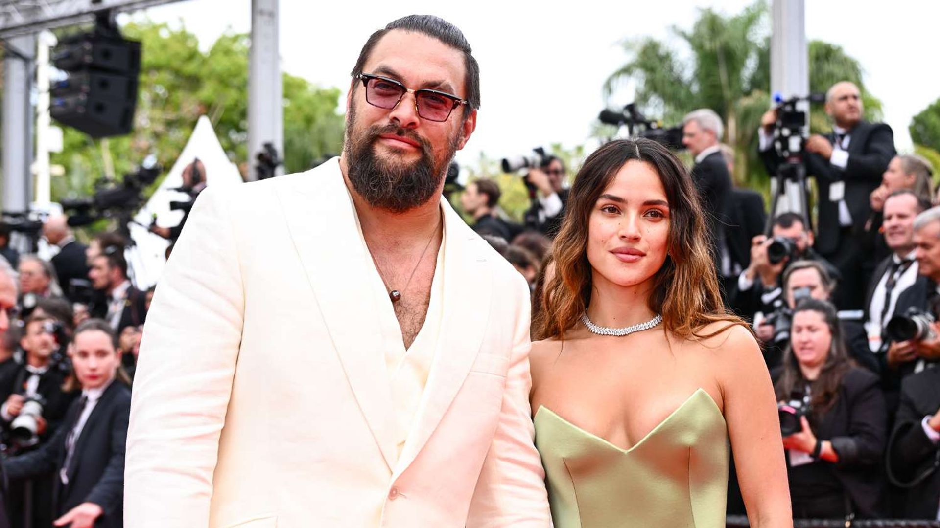 Jason Momoa de fato branco e Adria Arjona de vestido verde-oliva na passadeira vermelha