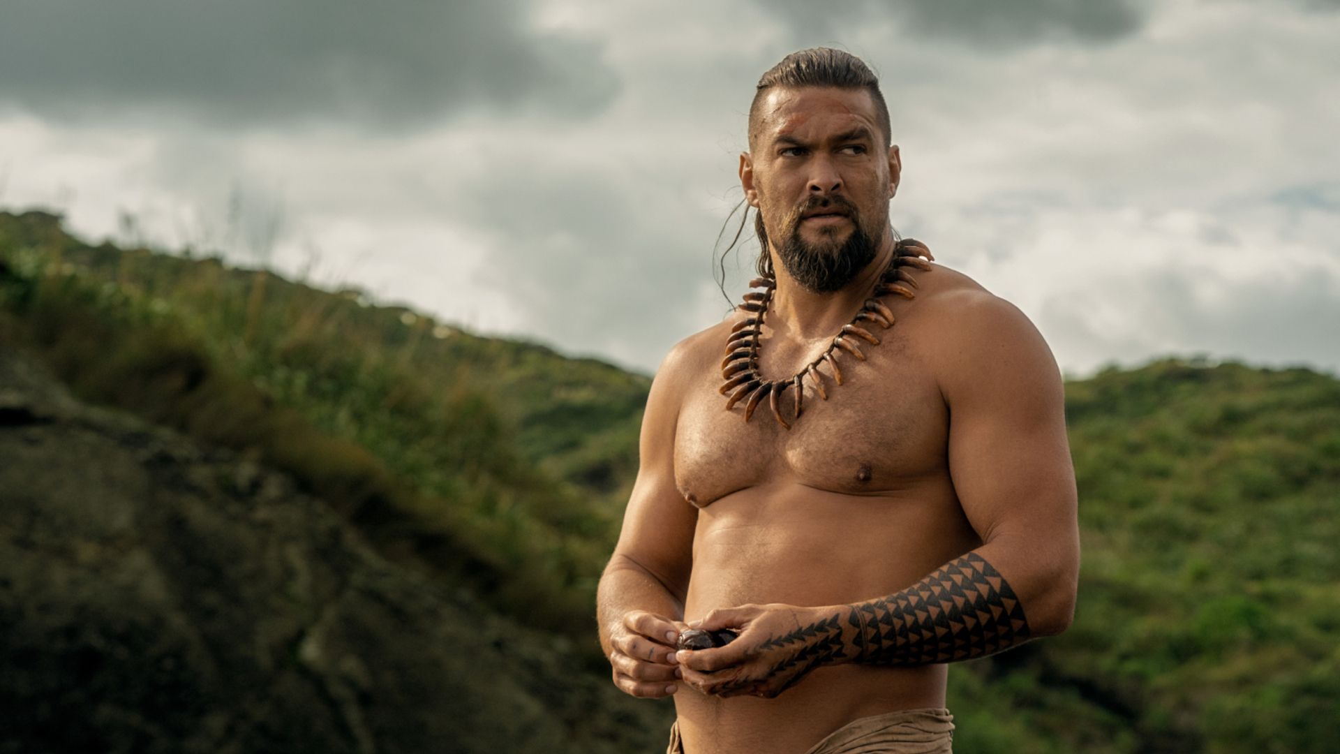 Jason Momoa sem camisa com decorações tradicionais havaianas e tatuagens contra um cenário montanhoso