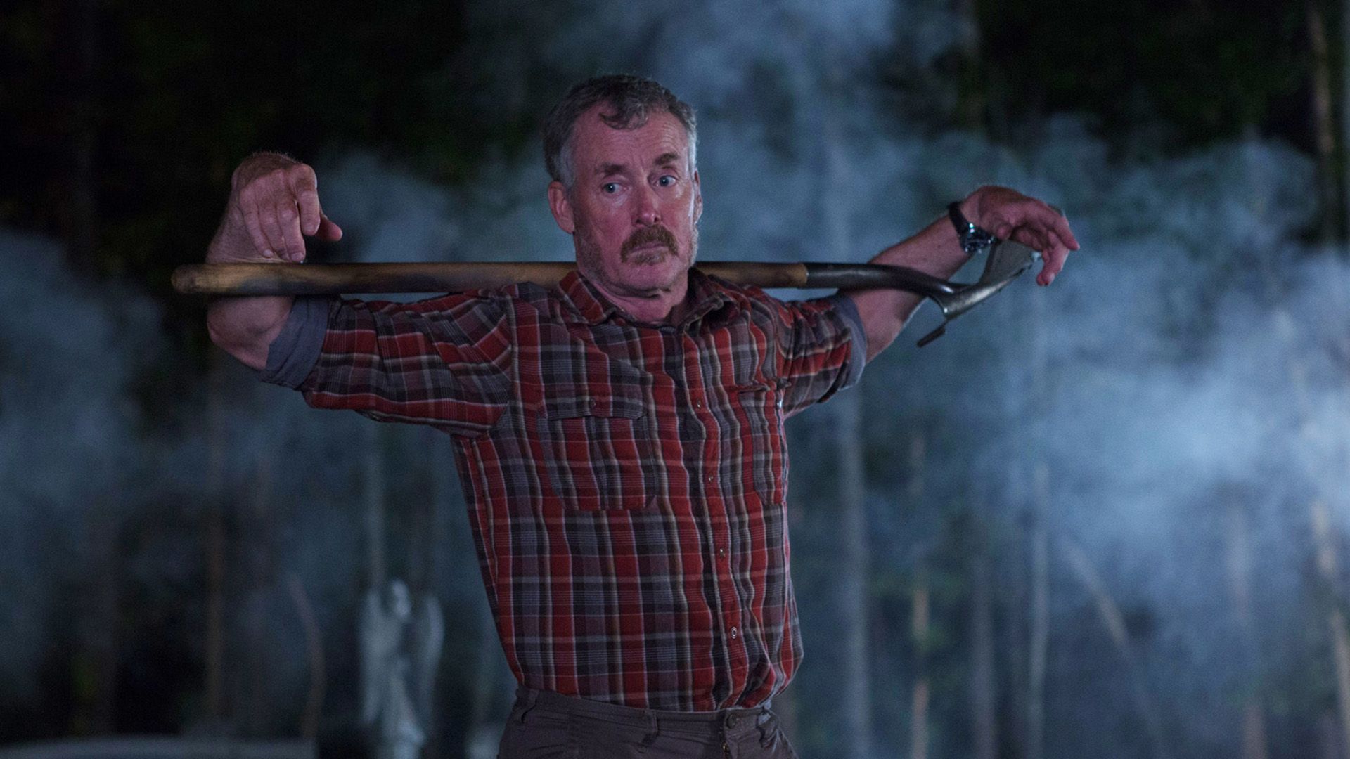 John C. McGinley numa camisa xadrez vermelha está na penumbra com uma pá aos ombros