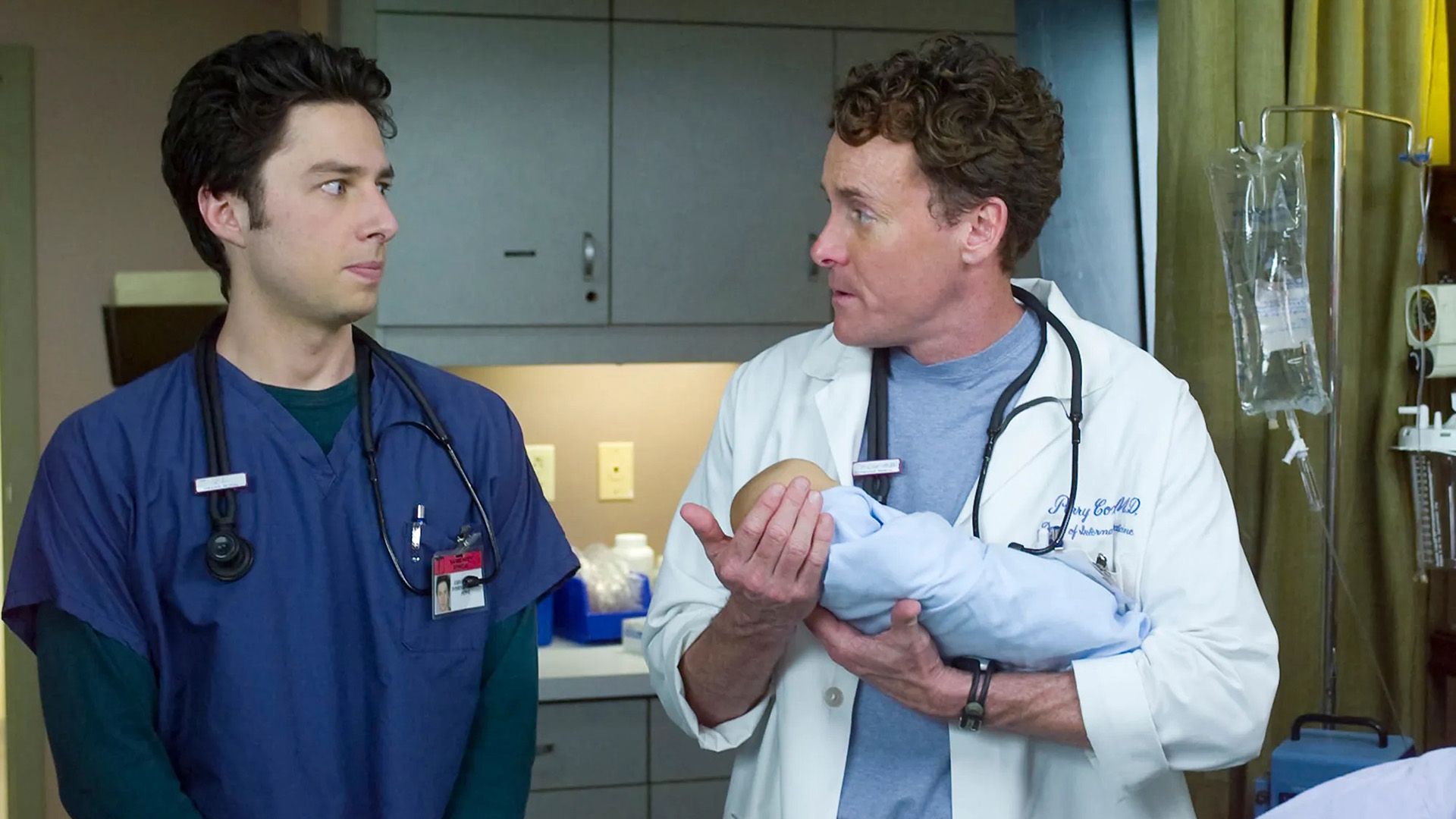 John C. McGinley numa bata branca a segurar um bebé, com Zach Braff ao lado vestido com uma bata azul